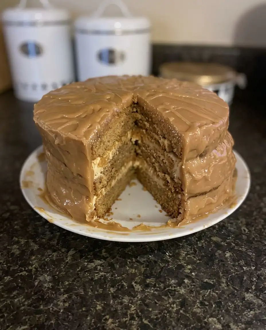 A person spreading thick caramel frosting on a yellow cake layer with an offset spatula.