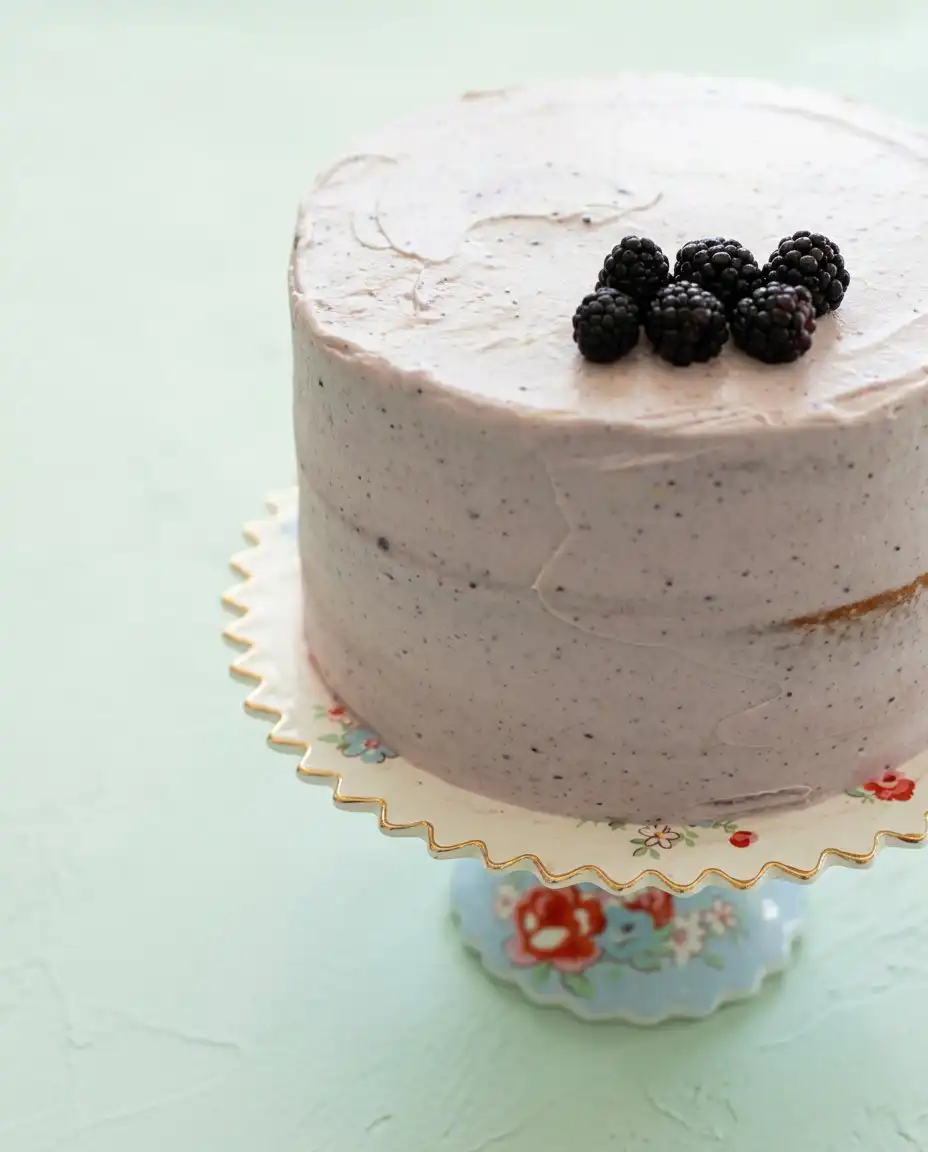 A slice of Blackberry Lemon Cake on a white plate next to a cup of coffee.