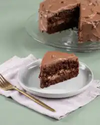 A thick slice of dairy-free chocolate zucchini cake sitting on a wooden counter with a glass dome.