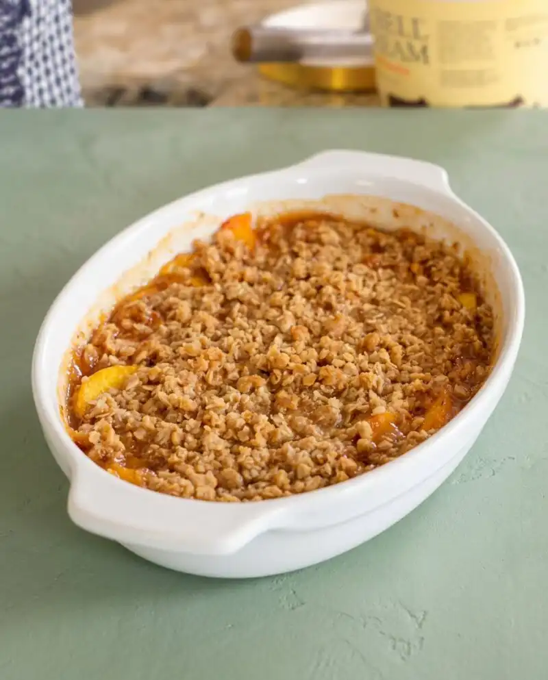 A golden brown Peach Crisp bubbling in a ceramic baking dish on a kitchen counter.
