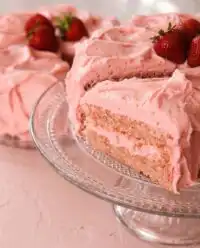 A beautiful two layer Strawberry Cake covered in pink cream cheese frosting on a white stand.
