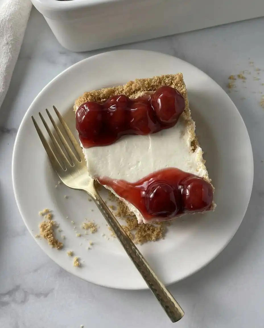 A spatula smoothing creamy white cheesecake filling over a buttery graham cracker crust in a 13x9 pan.