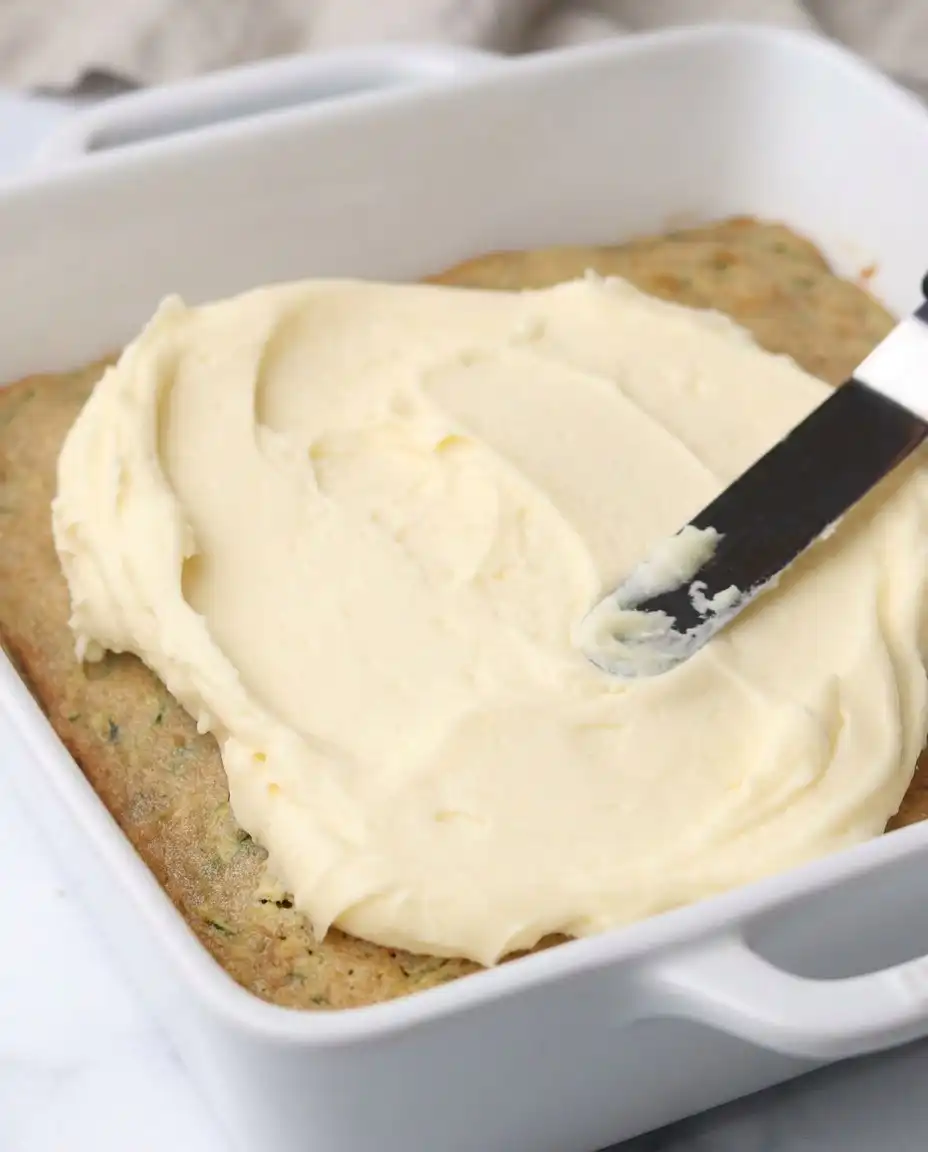 A hand spreading cream cheese frosting over a pan of baked Zucchini Bars.