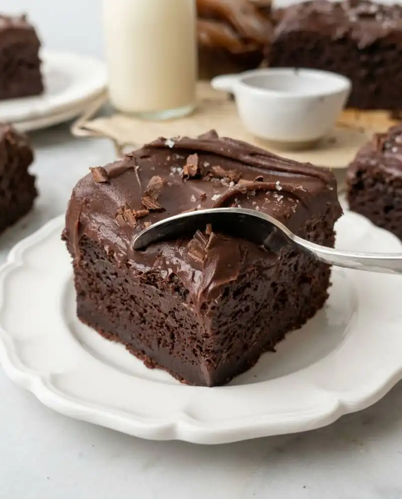 A moist Vegan Chocolate Zucchini Cake sitting under a glass dome on a wooden kitchen counter.