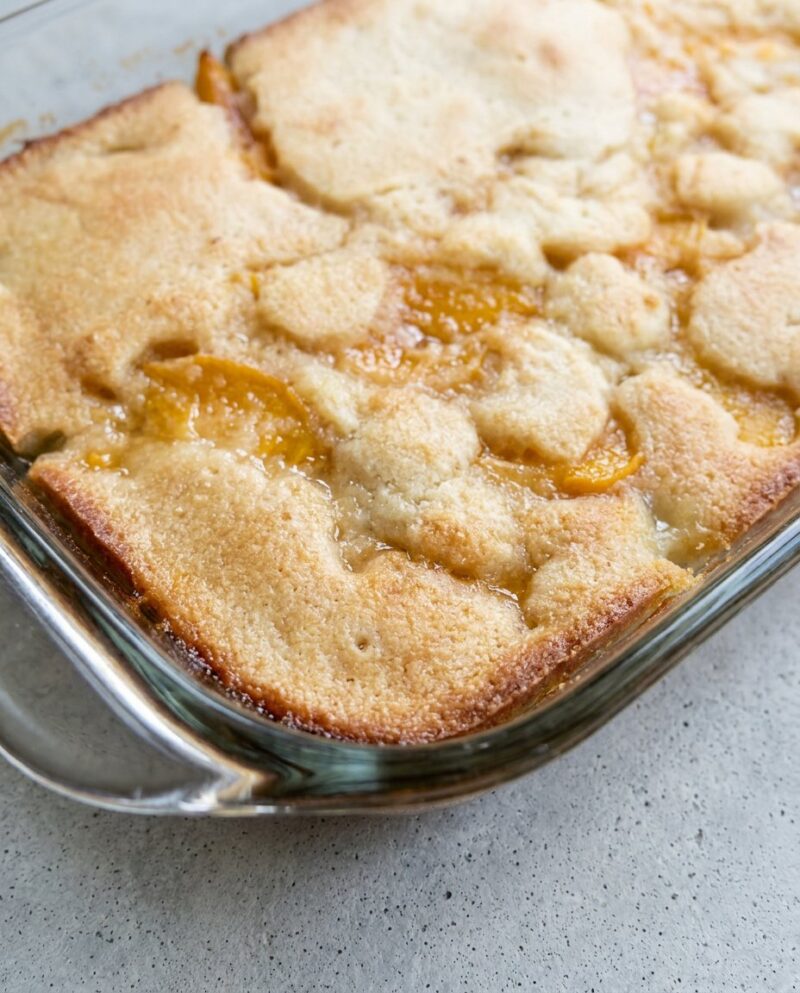 A golden brown Peach Cobbler with Canned Peaches served in a white ceramic baking dish on a counter.