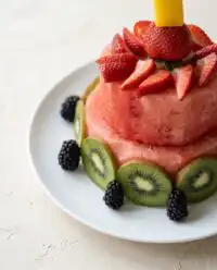 A tiered Baby's First Watermelon Yogurt Cake decorated with fresh strawberries and blueberries on a wooden serving board