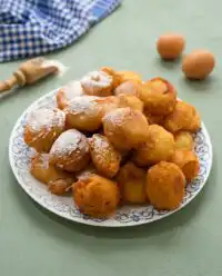 A pile of golden brown classic funnel cake bites covered in a dusting of powdered sugar on a plate.
