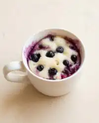 A warm and fluffy Easy Blueberry Mug Cake topped with juicy fresh blueberries in a white ceramic mug.