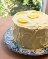 A beautiful three layer Lemon Blueberry Layer Cake with Cream Cheese Frosting resting under a glass dome on a wooden counter.