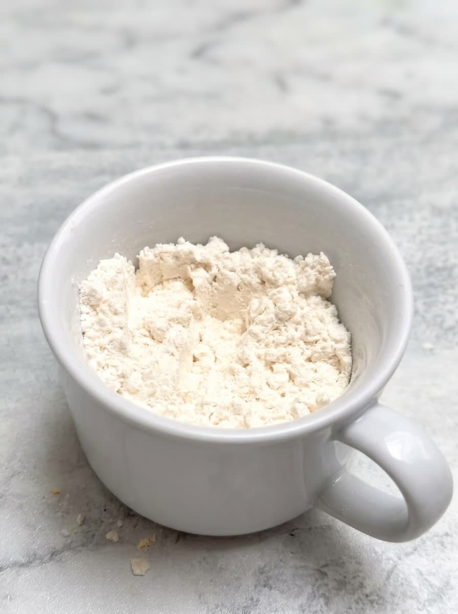 1-Minute Vanilla Mug Cake 14 Measured dry and wet ingredients for making a quick 1-Minute Vanilla Mug Cake on a rustic wooden counter