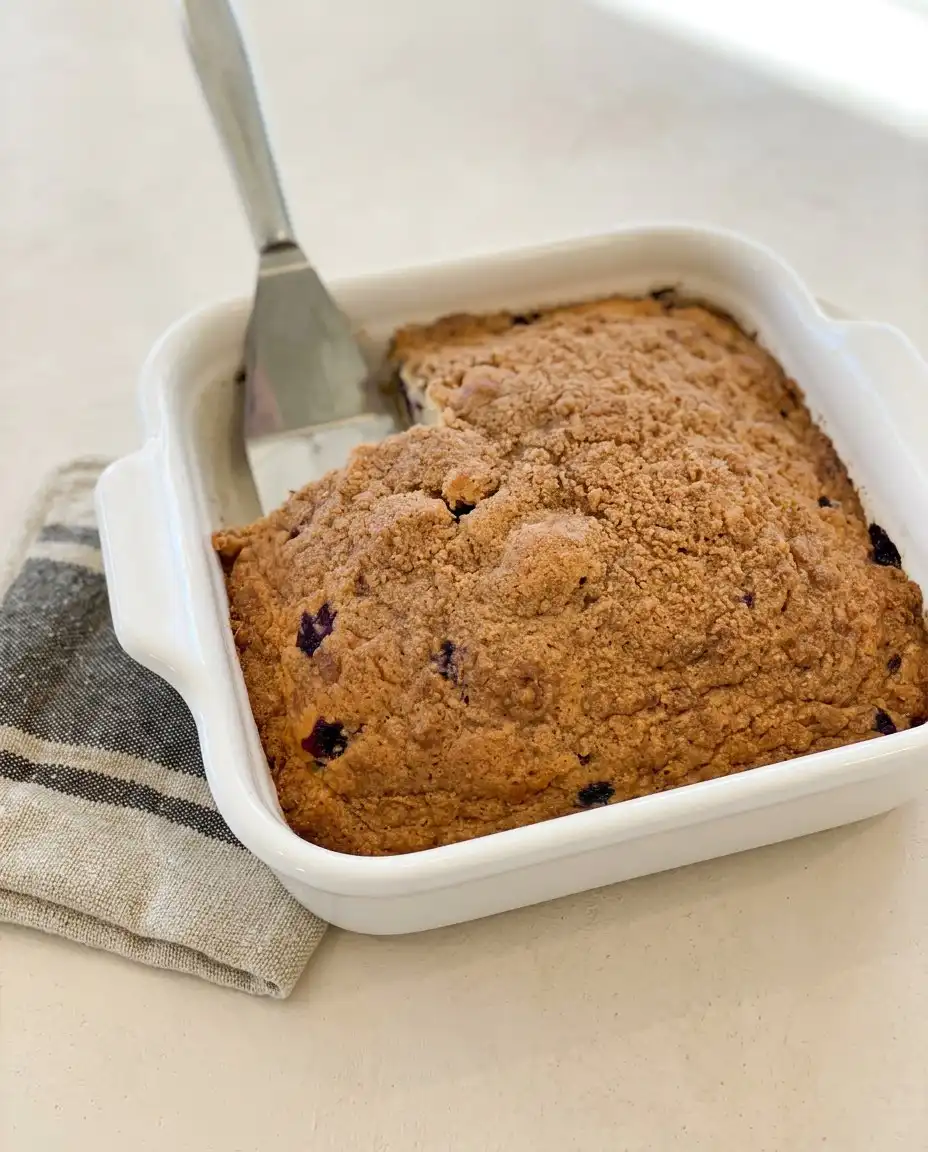A whole square blueberry coffee cake sliced and served with a cup of hot coffee.