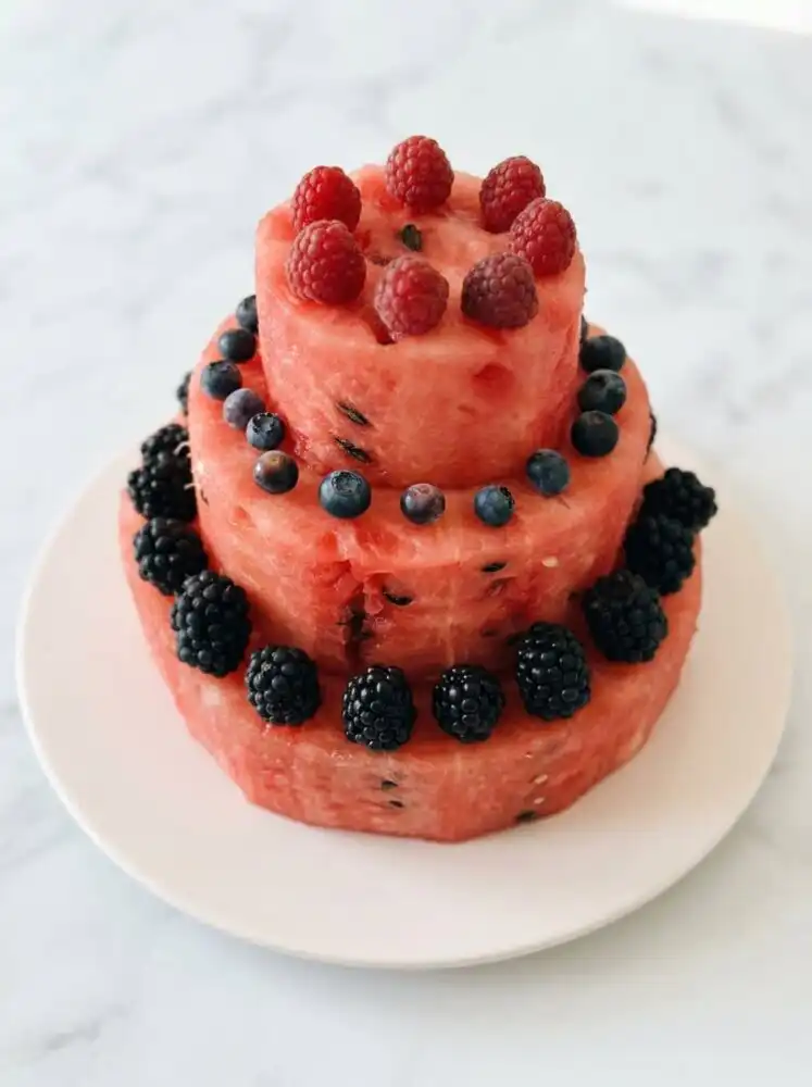 A beautifully sliced Watermelon and Berry Layer Cake topped with fresh peaches and blueberries on a wooden table.
